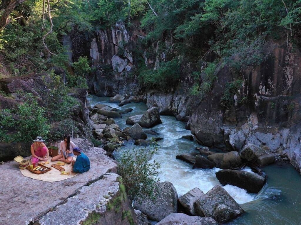 a group of people sitting on a blanket on a rock ledge by a river